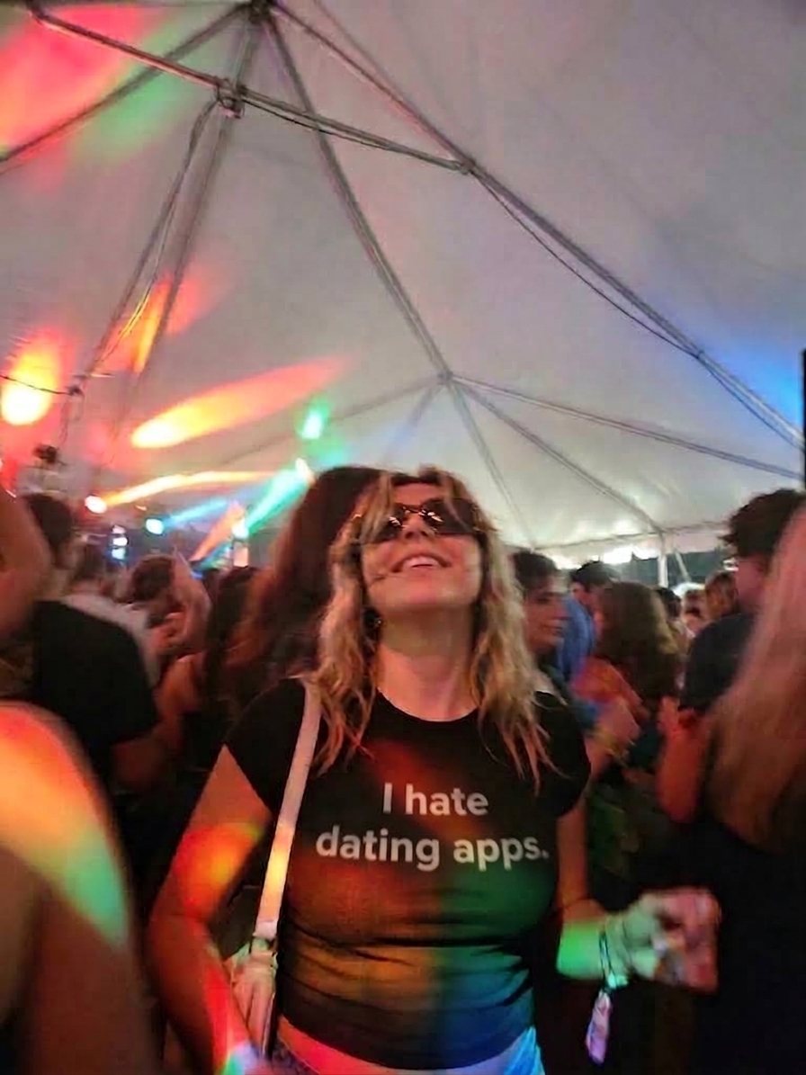 Woman in a crowded festival tent wearing a black Sparkbite baby tee that reads "I hate dating apps."